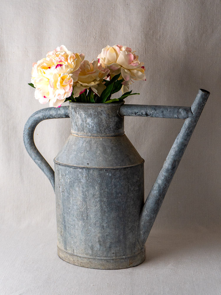 Antique French zinc long-spout watering can planter with pink and cream flowers on neutral fabric.
