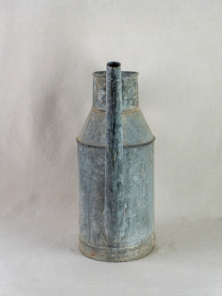 Antique French zinc arrosoir with cylindrical body and long spout on neutral fabric background.