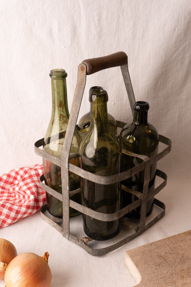 Antique French zinc and wood four-bottle wine carrier with green glass bottles, wooden handle, on light fabric.