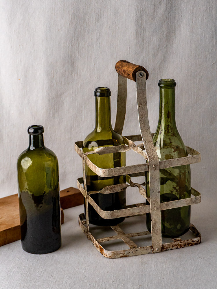 Three empty green glass bottles, two in an antique painted zinc French wine carrier with wooden handle.