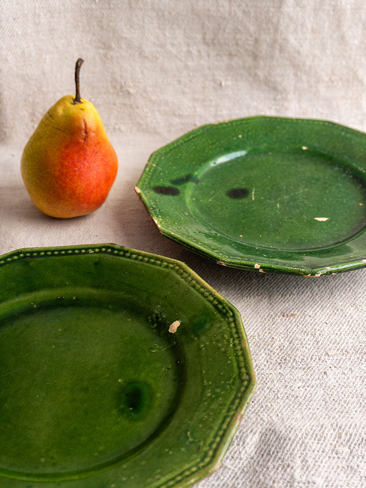 Pair of antique French green glazed plates with chipped edges, shown beside a ripe pear on textured beige fabric.