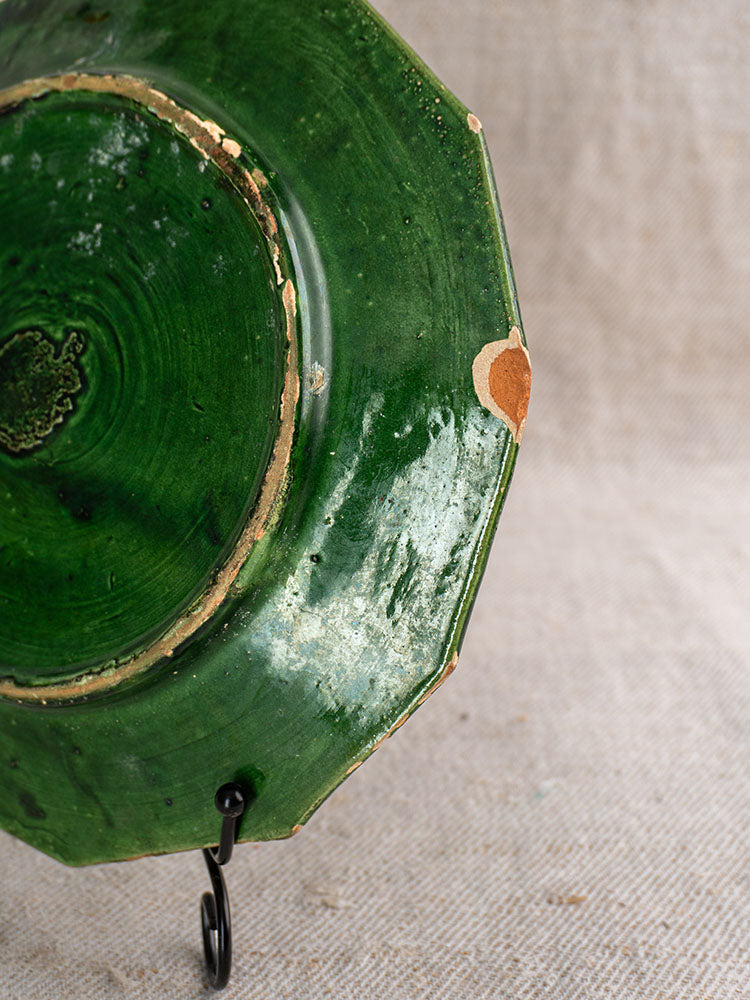 Antique French green glazed plate with chipped edges, exposed clay, on black metal stand and beige fabric.