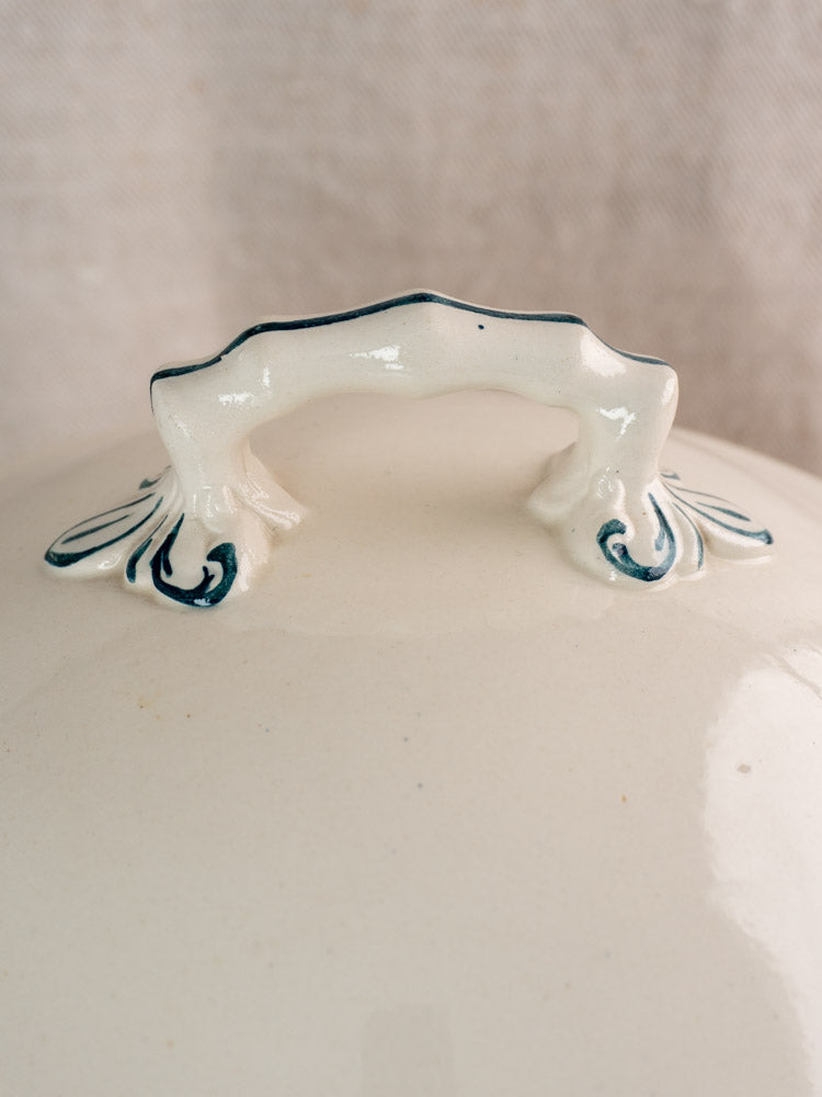Ornate arched lid handle with blue accents on antique French Longwy ironstone Capucine tureen, neutral fabric backdrop.