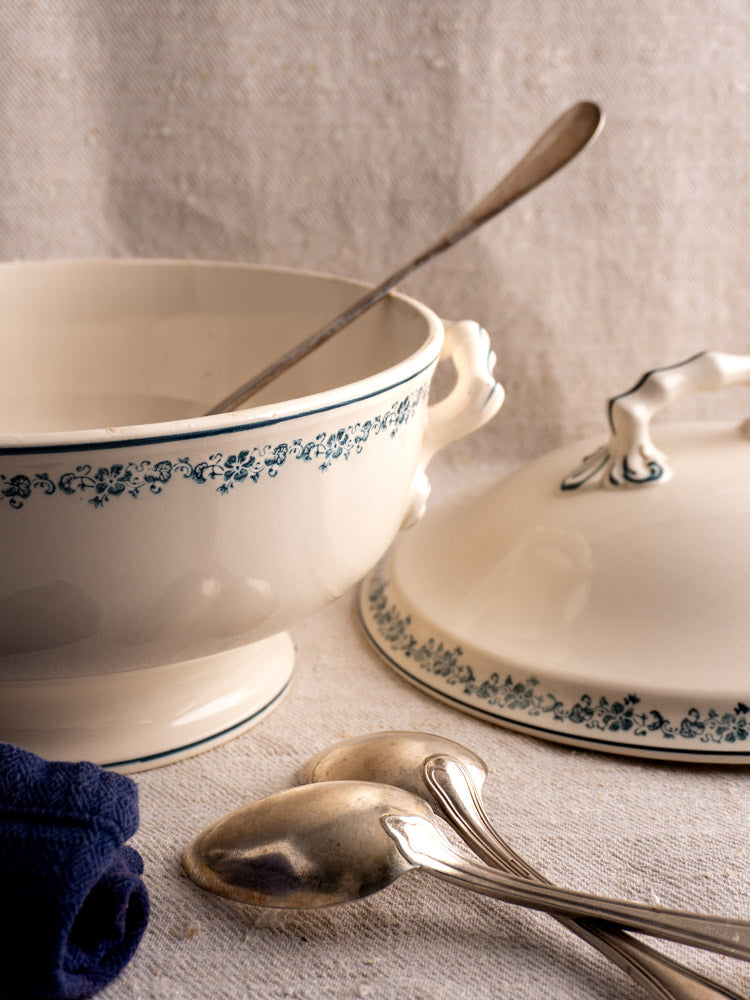 Antique French Longwy ironstone soup tureen with Capucine blue floral trim, lid, and silver spoons on textured cloth.