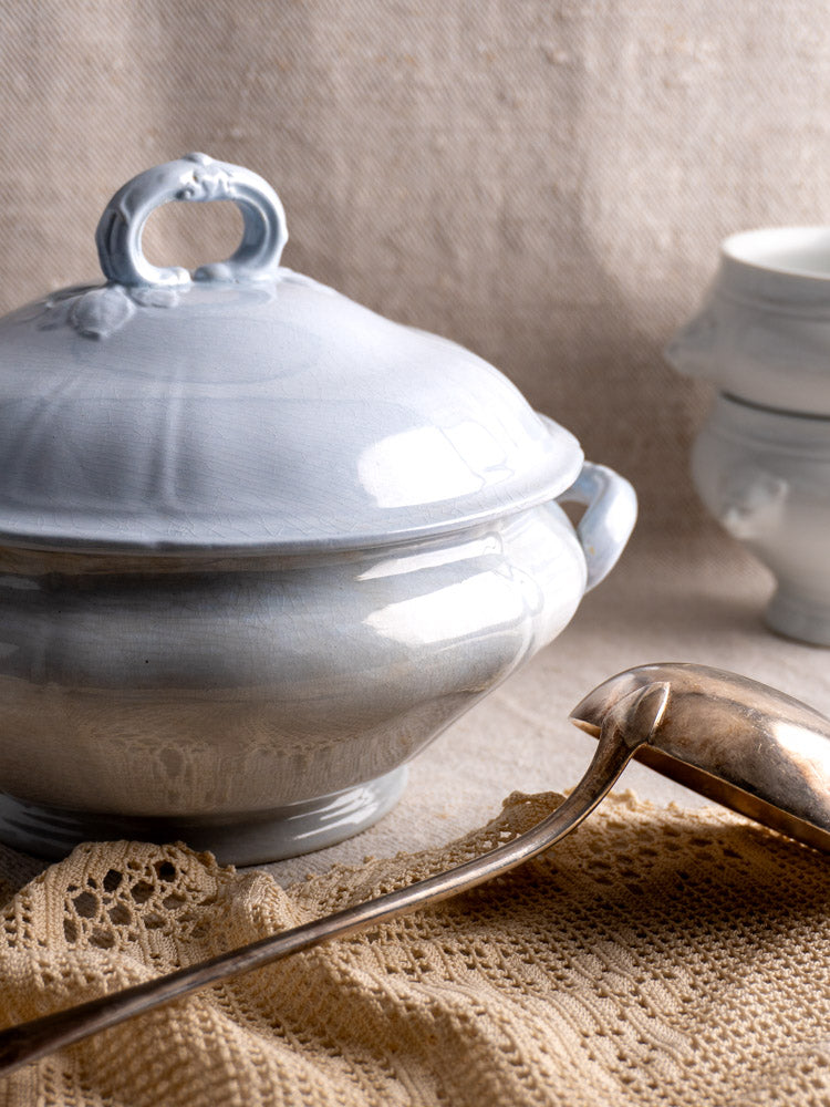 Antique Villeroy and Boch Mettlach tureen with soft blue glaze and lid, on lace cloth beside silver ladle.