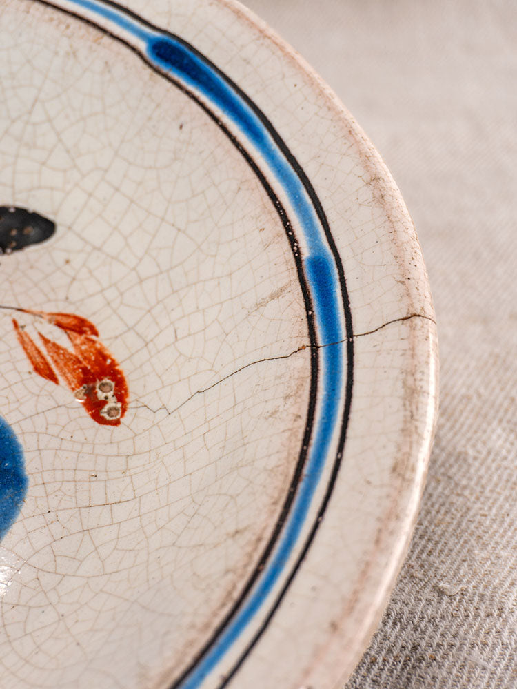 Antique French faience plate with blue rim, orange fish, visible crack, age signs on textured light fabric.