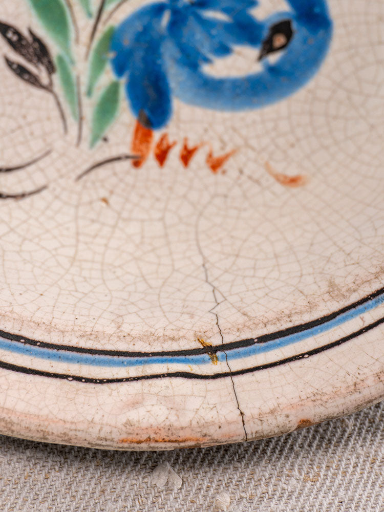 Antique French faience plate with blue floral motifs, crackle glaze, and rim cracks on textured fabric background.