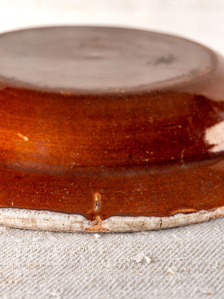 Antique French faience plate, Cul Noir style, brown underside with chipped edges, on light textured surface.