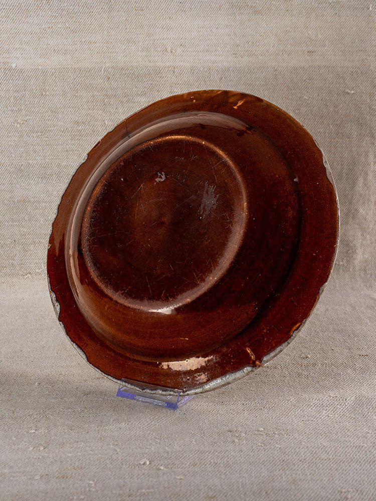 19th-century French faience plate, glossy brown with wear, displayed on clear stand, textured beige fabric backdrop.