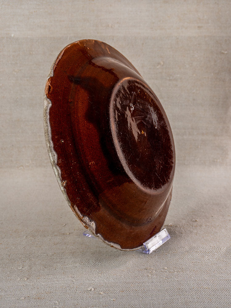 Antique French faience plate with rustic uneven edges and glossy brown center on clear stand, beige textured background.