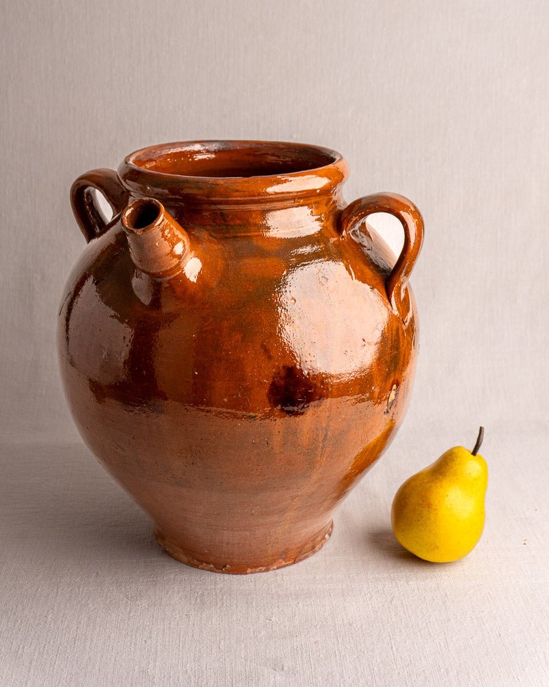 Antique French glazed terracotta oil jar with three handles and brown finish beside a yellow pear.
