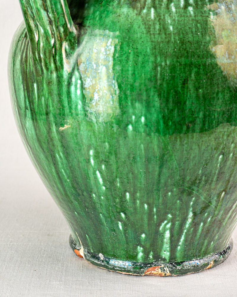 Antique French green glazed earthenware cruche with worn glossy texture and brown chips, on a neutral background.