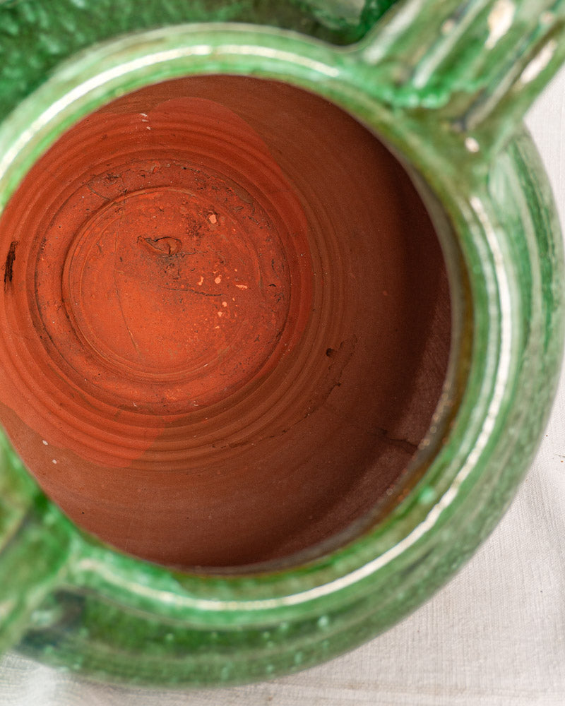 Top view of antique French green glazed earthenware cruche with reddish-brown interior on light fabric surface.