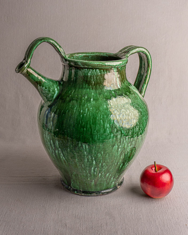19th-century French green glazed earthenware cruche with two handles beside a red apple on gray background.