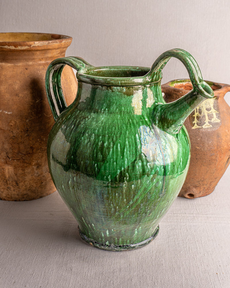 19th-century French green glazed earthenware cruche with two handles and spout, in front of brown pottery.