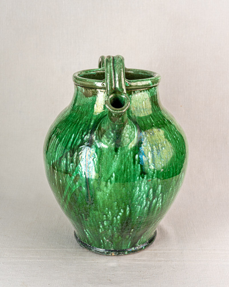 19th-century French green glazed earthenware cruche with round body, two spouts, and handle on light textured surface.