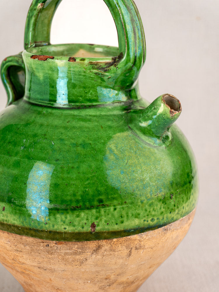 Antique French green-glazed terracotta gargoulette with rustic texture, spout, handle, and chipped rim on a plain background.