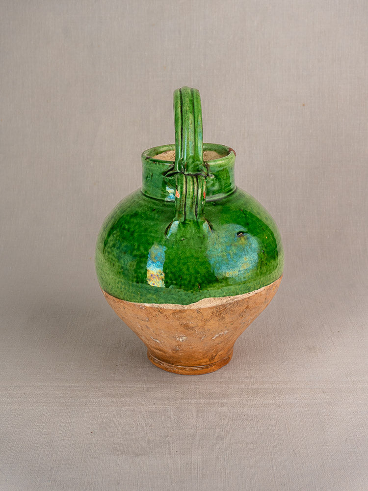 Antique French gargoulette cruche in green glazed terracotta with clay lower half and curved neck handle, on a light background.