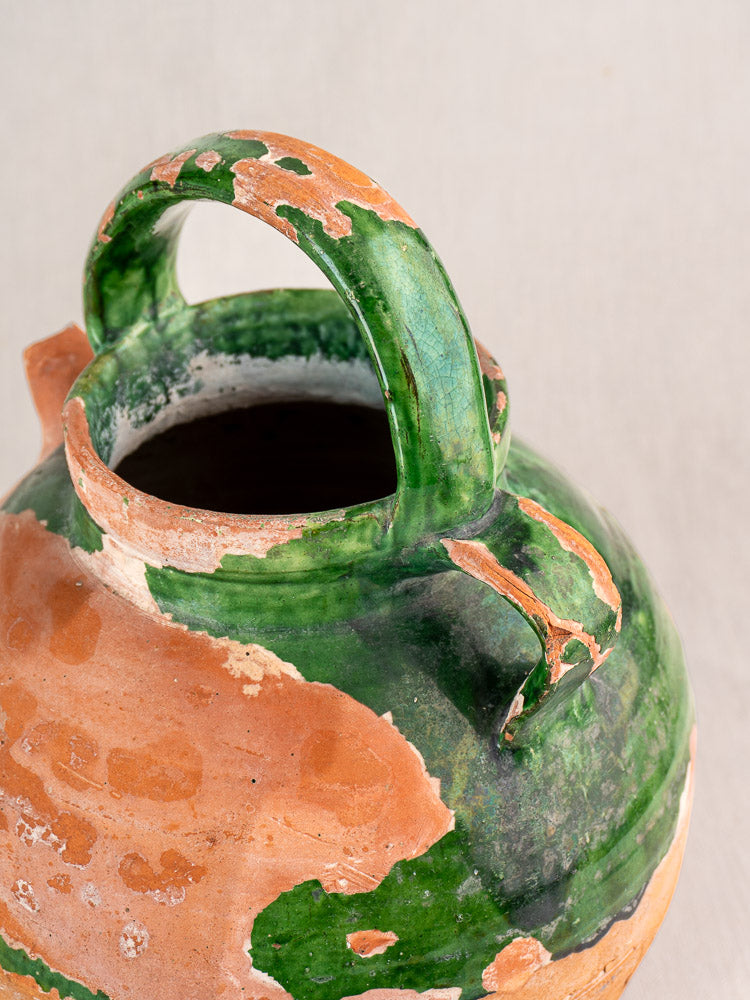 Antique French green glazed earthenware gargoulette jug with handle, terracotta chips; light fabric background.