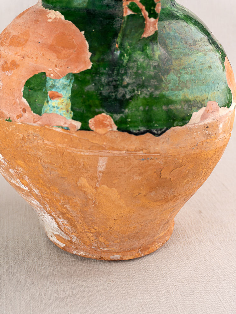 Antique French green-glazed earthenware gargoulette with chipped glaze and exposed terracotta on neutral surface.