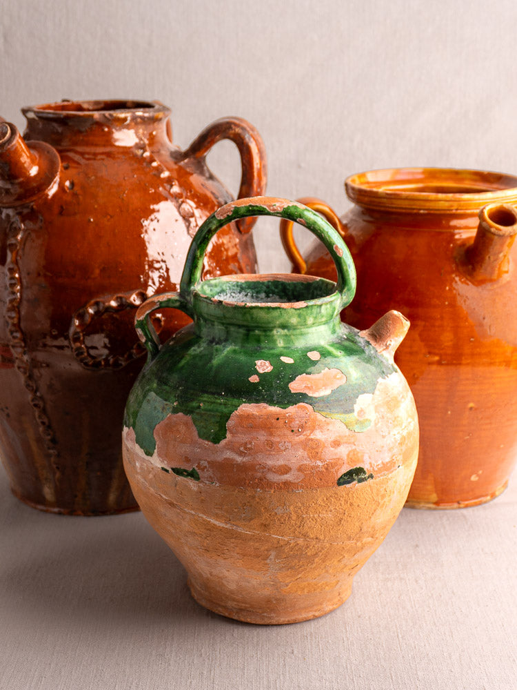 Antique French green-glazed earthenware gargoulette and two reddish-brown ceramic jugs on light fabric, neutral background.