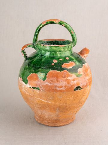 Antique French green glazed earthenware gargoulette water jug with two handles, unglazed brown base, on a light grey surface.