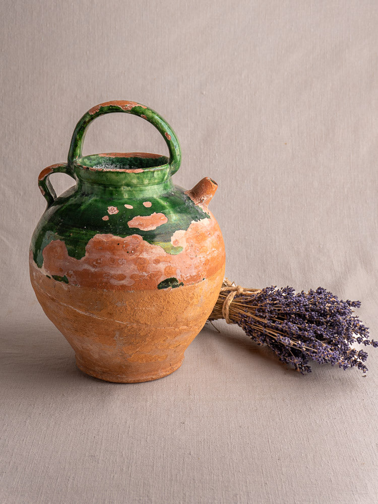 Antique French green glazed earthenware gargoulette water jug with large handle beside dried lavender on neutral fabric.