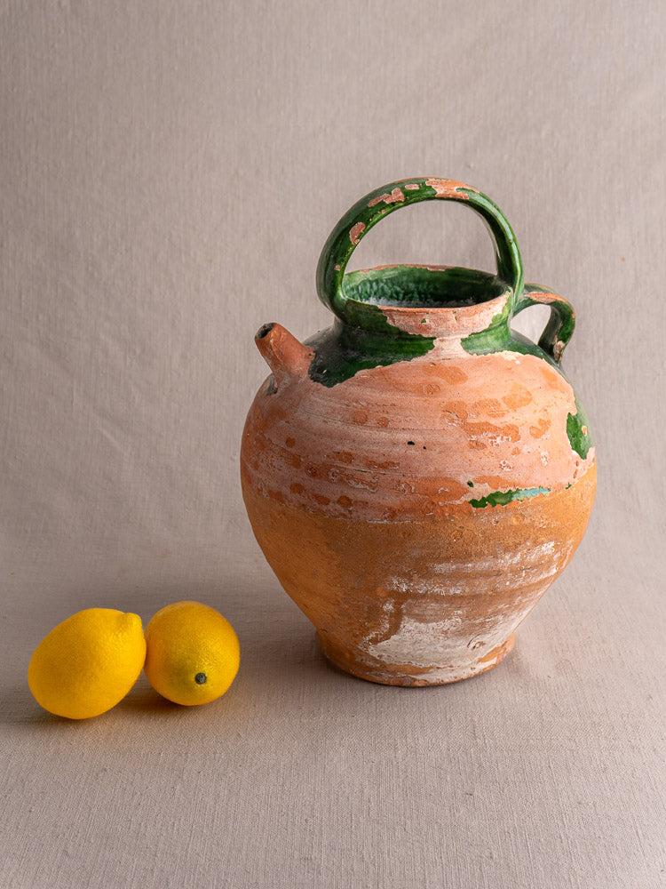 Antique French green-glazed earthenware gargoulette water jug with two yellow lemons on a light beige surface.