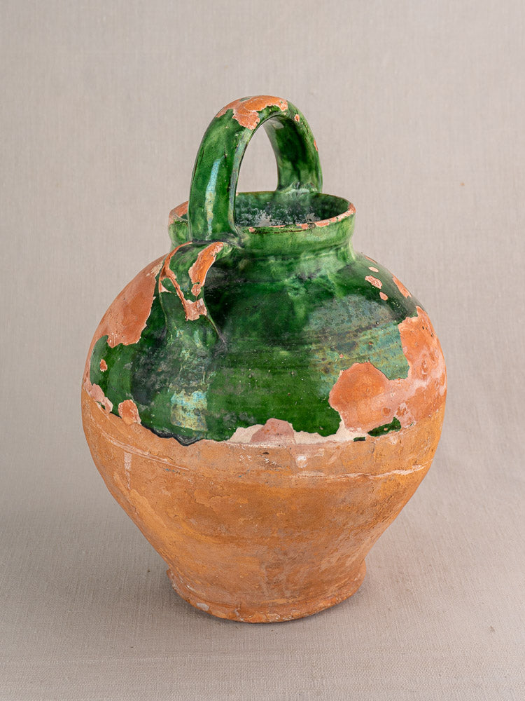 Antique French green glazed earthenware gargoulette water jug with curved handle, terracotta wear, neutral backdrop.