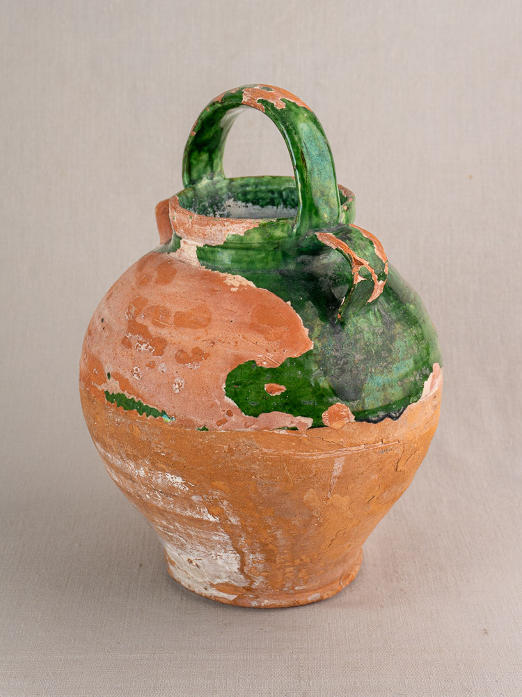 Antique French green glazed earthenware gargoulette with single handle and terracotta accents on neutral fabric background.