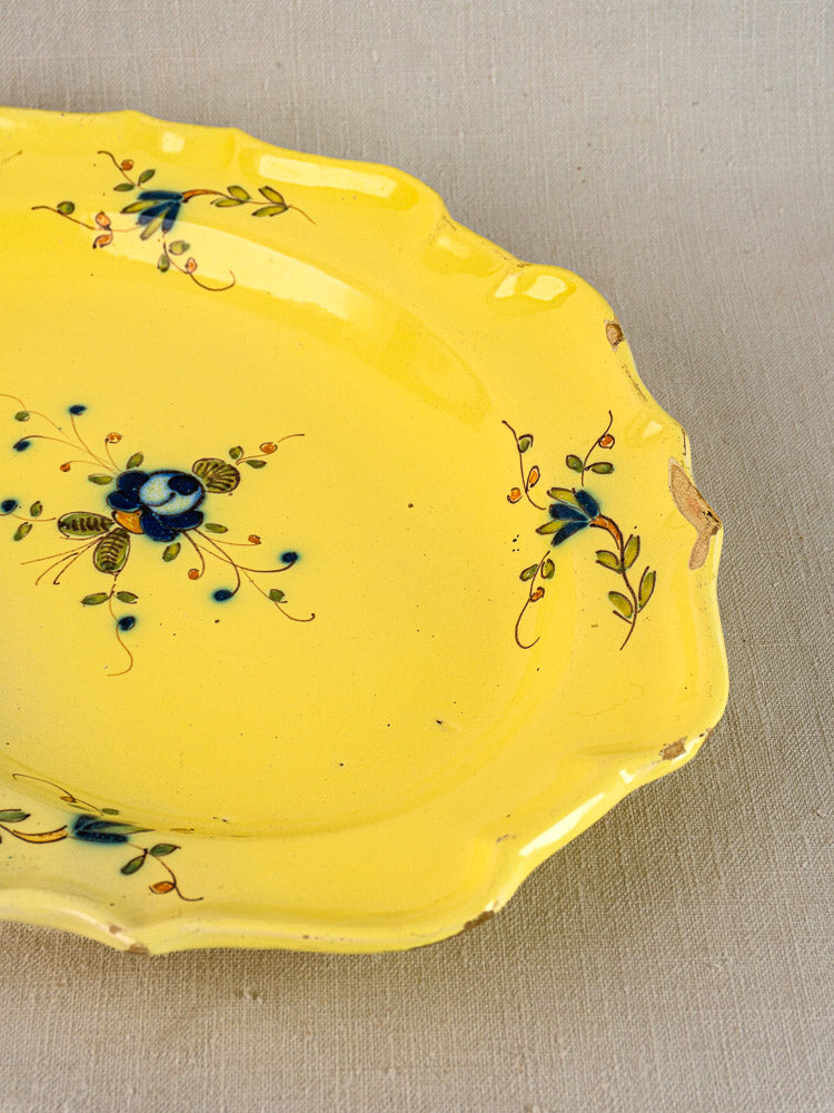 Antique French faience platter with yellow glaze, blue floral motifs, scalloped edge, rim chips; on beige fabric.