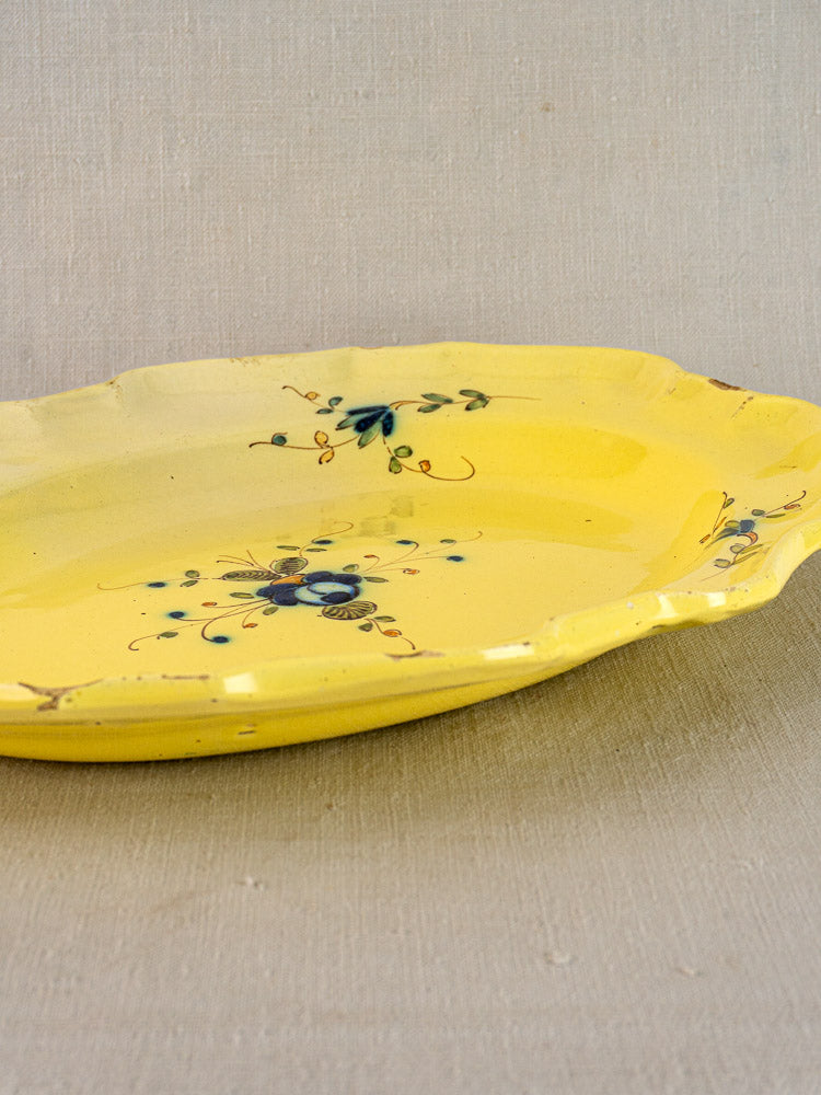 French faience platter with yellow glaze, scalloped edges, and hand-painted florals on earthenware, on neutral fabric background.