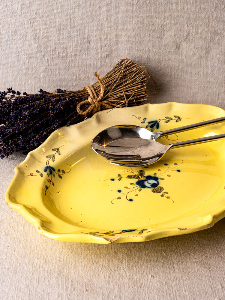 Antique French faience platter with yellow glaze, blue floral motifs, and silver serving utensils beside dried lavender.