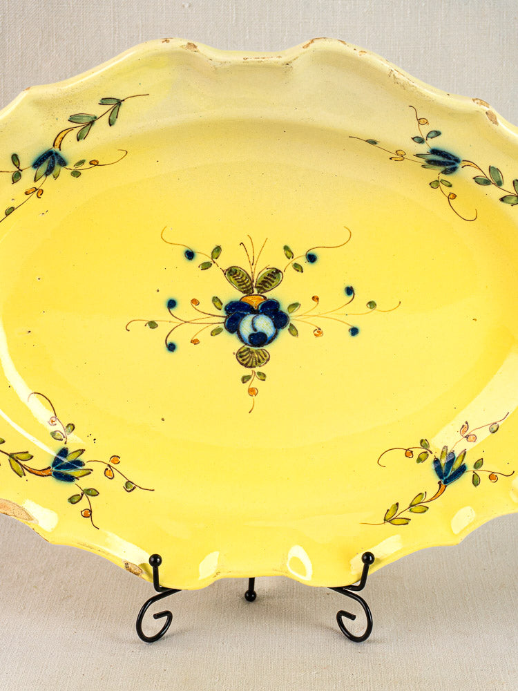Antique French faience platter with yellow glaze, blue-green floral motifs, and scalloped edges on black metal stand.