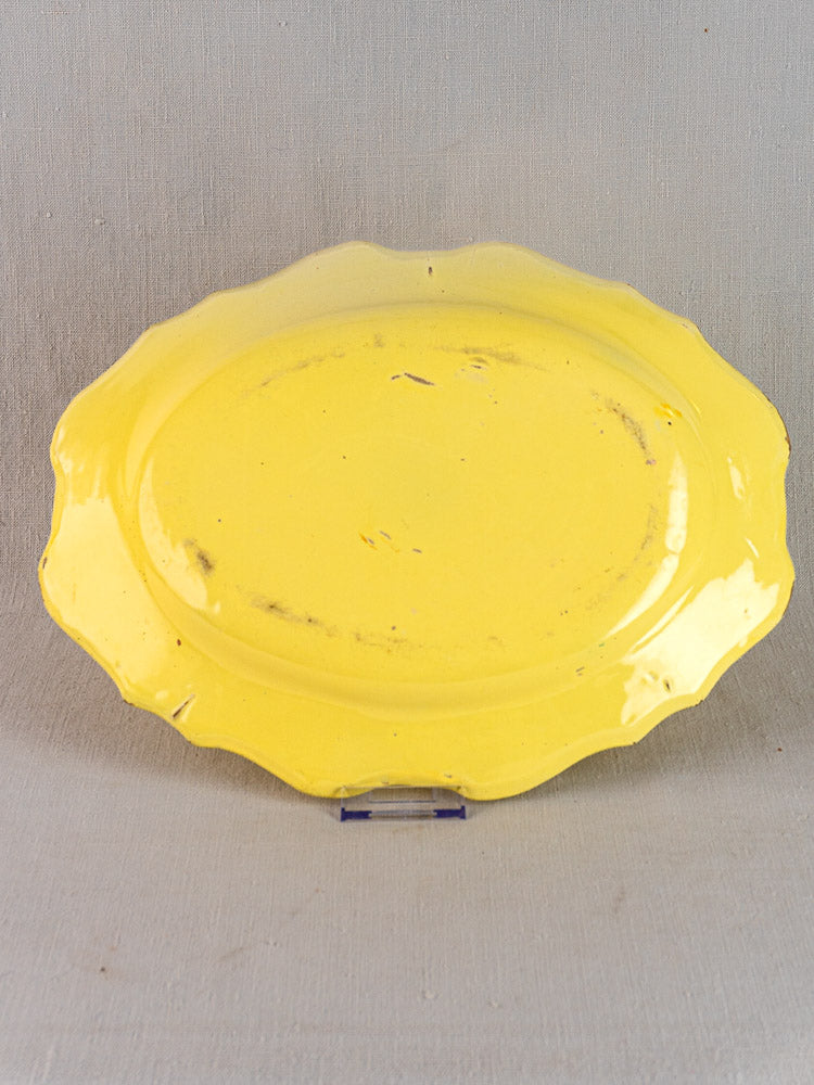Antique French faience oval platter with yellow glaze and scalloped edges, light scratches; upright on textured gray backdrop.