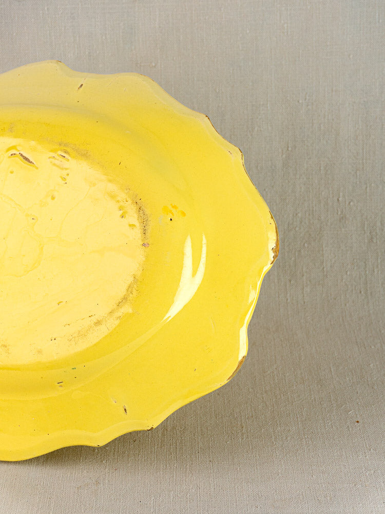 Antique French faience platter with bright yellow glaze and scalloped edge, partially on light gray textured surface.