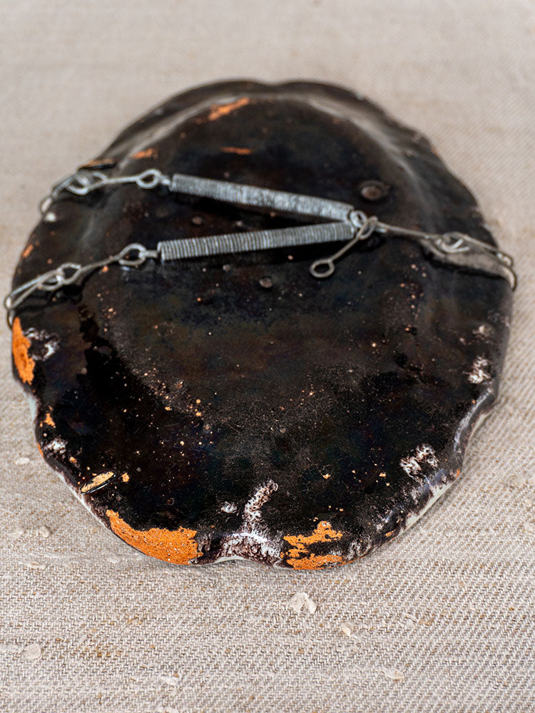 Antique French faience platter with irregular dark ceramic form, metal wire accents, on neutral fabric background.