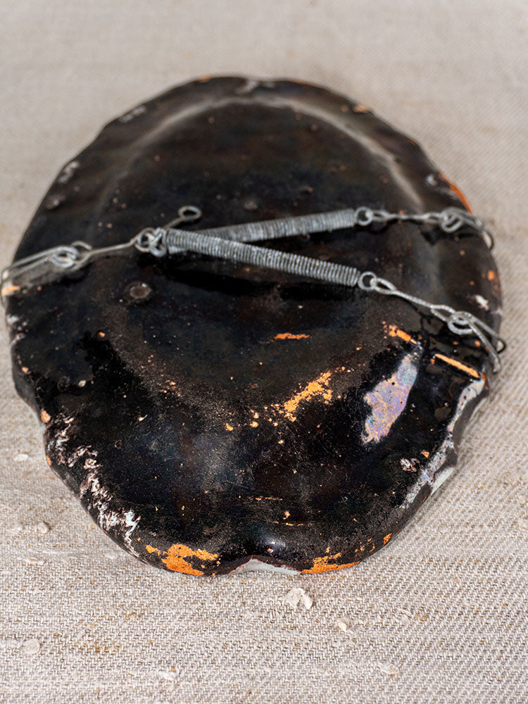Dark oval ceramic mask with metal wire and springs on textured beige fabric, inspired by antique French faience artistry.