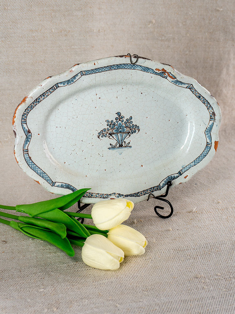 Antique French 18th-century faience platter with blue floral motif, worn edges on metal stand, white tulips on beige surface.