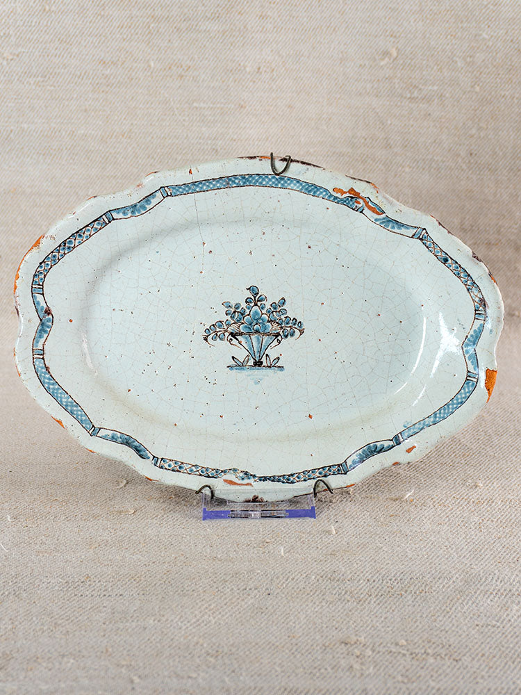 18th-century French faience platter with blue floral center, patterned border, edge wear, on stand against beige fabric backdrop.