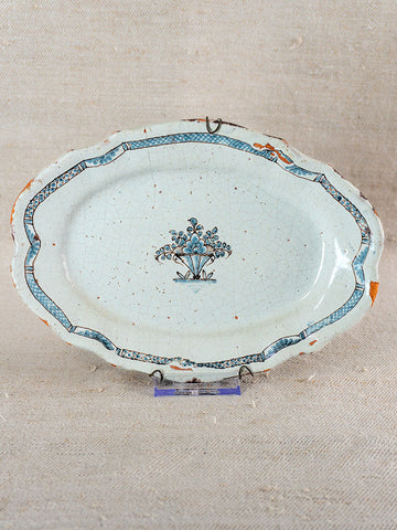 18th-century French faience platter with blue floral center, patterned border, edge wear, on stand against beige fabric backdrop.