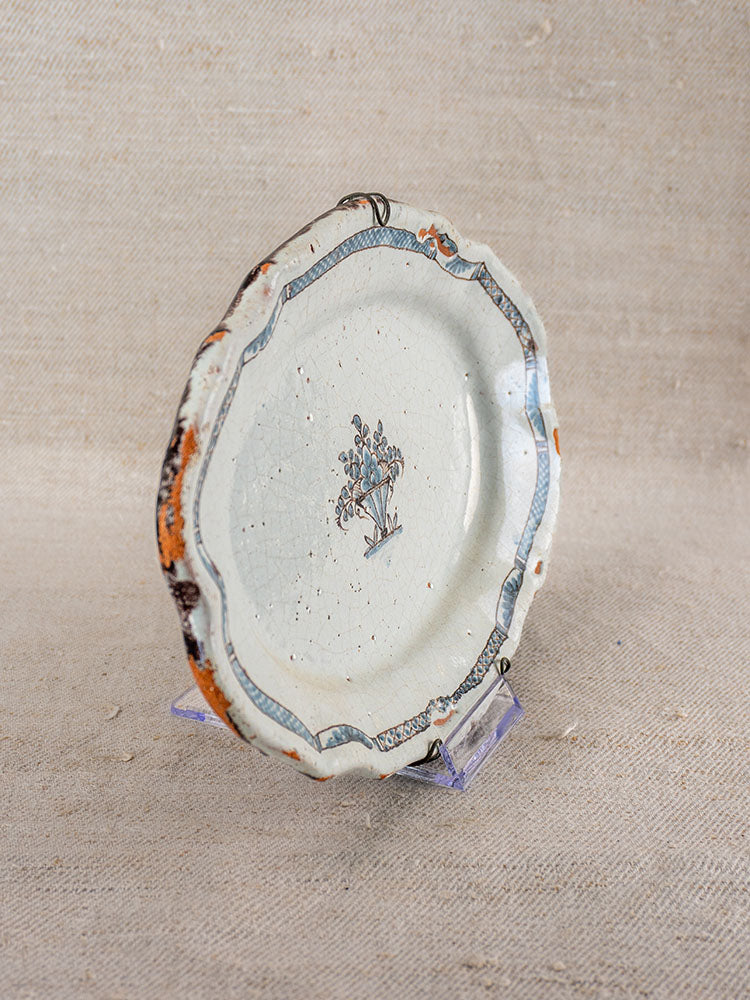 18th-century French faience platter with scalloped edge, blue and brown floral motifs on clear stand, burlap backdrop.