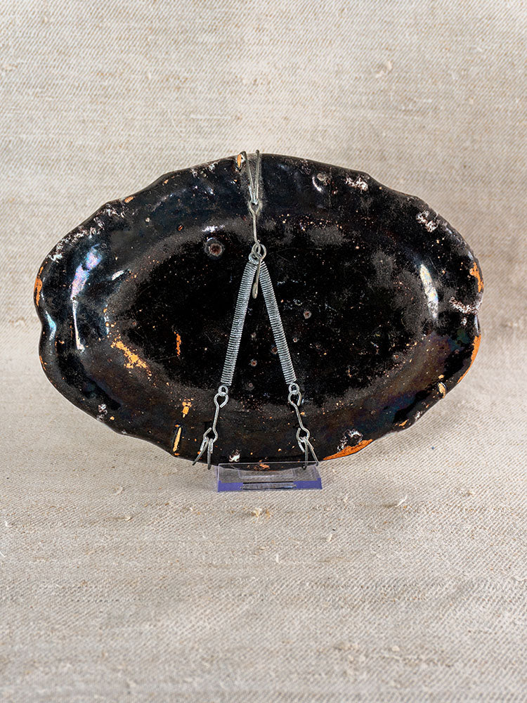 Antique 18th-century French faience platter, Cul Noir, dark glaze, oval with uneven rim on beige fabric and metal stand.