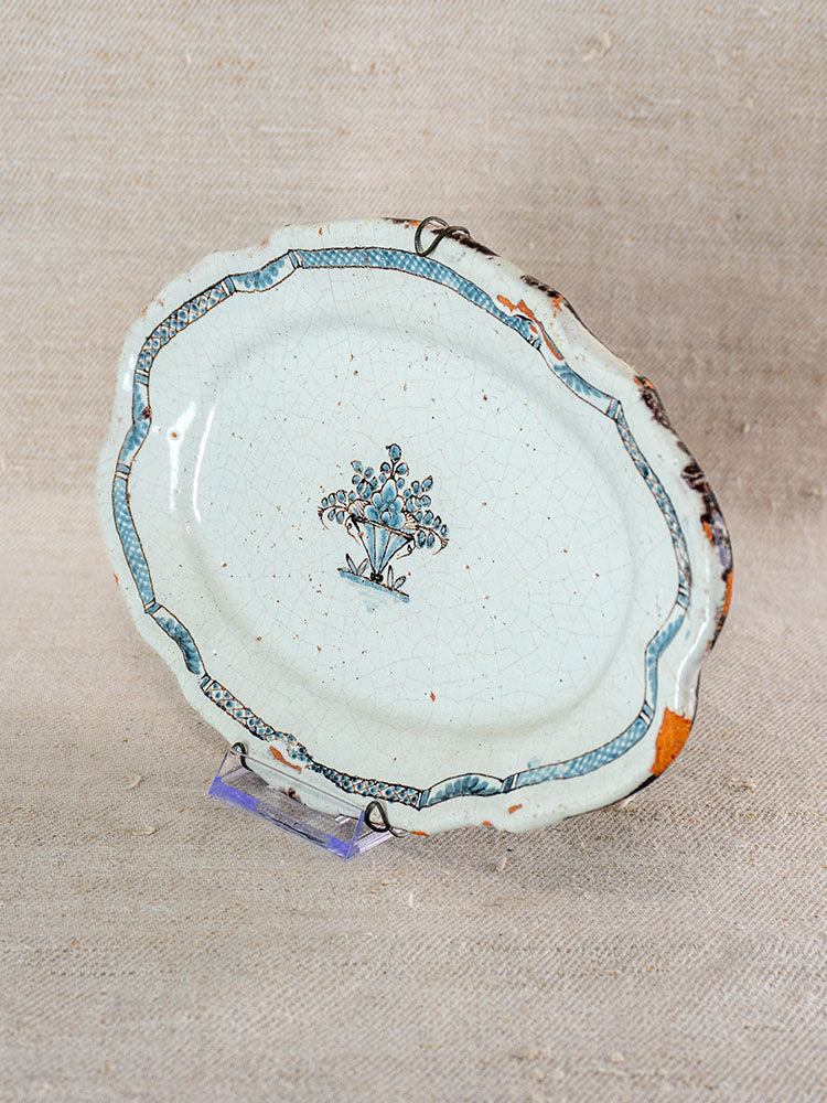 18th-century French faience platter with blue floral motif and border, visible chips and cracks, on fabric surface.