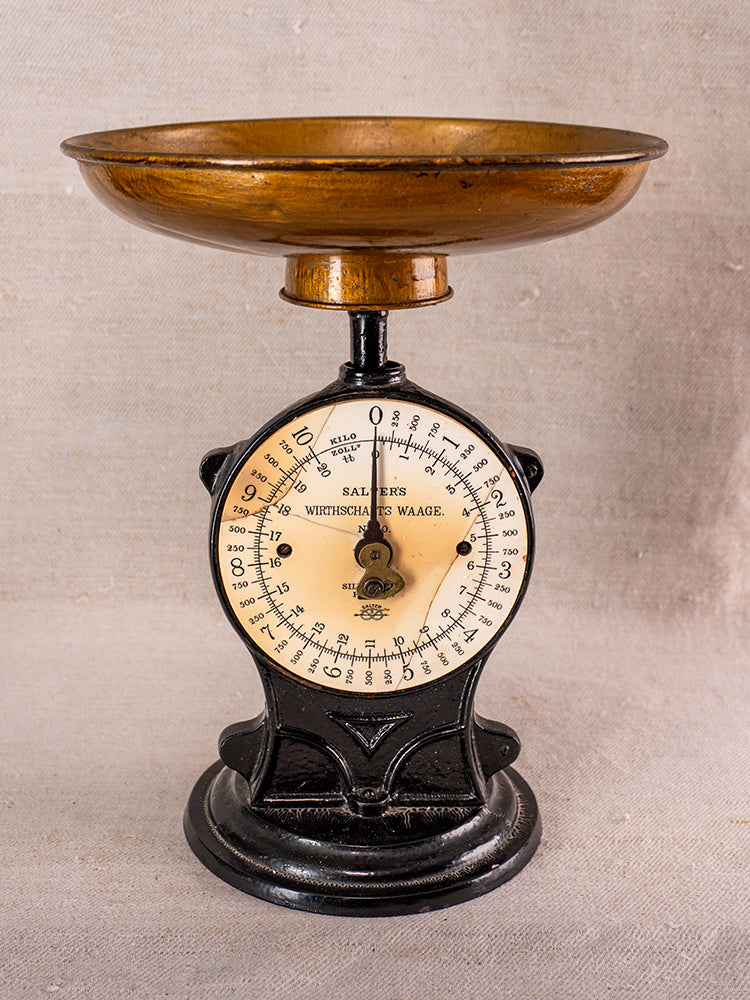 Antique Salter cast iron kitchen scale with black and gold dial, brass pan, on neutral fabric background.