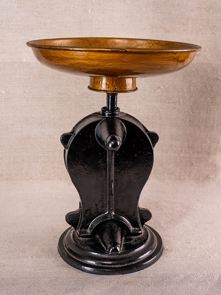 Decorative pedestal bowl with wooden top and ornate black metal base, inspired by antique kitchen scale, on beige fabric.
