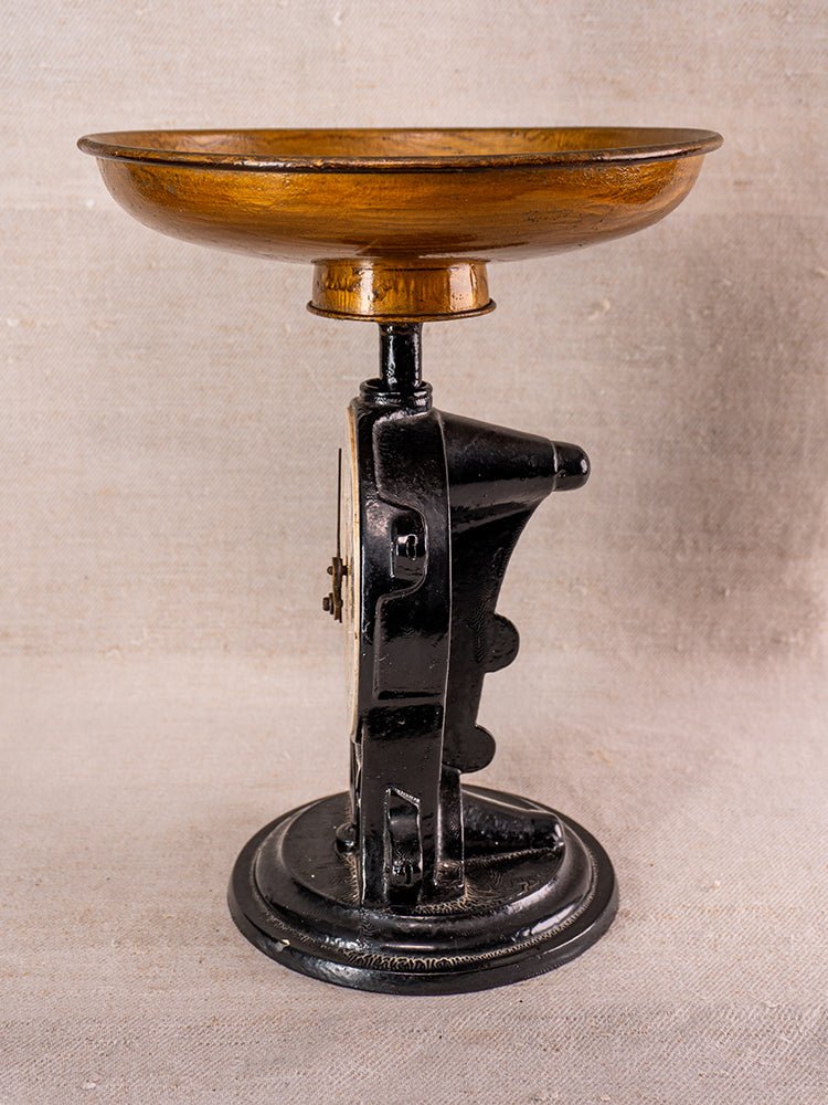 Antique Salter cast iron kitchen scale with black body and round brass tray on neutral fabric background.