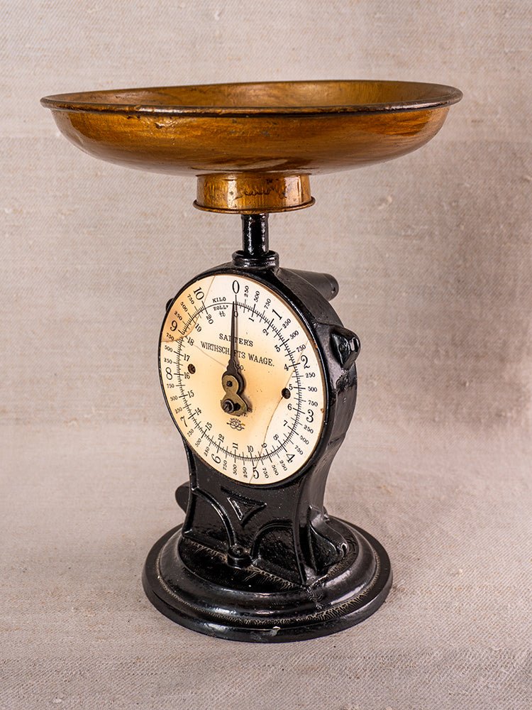 Antique cast iron Salter kitchen scale with black and brass finish, round dial, on neutral fabric background.