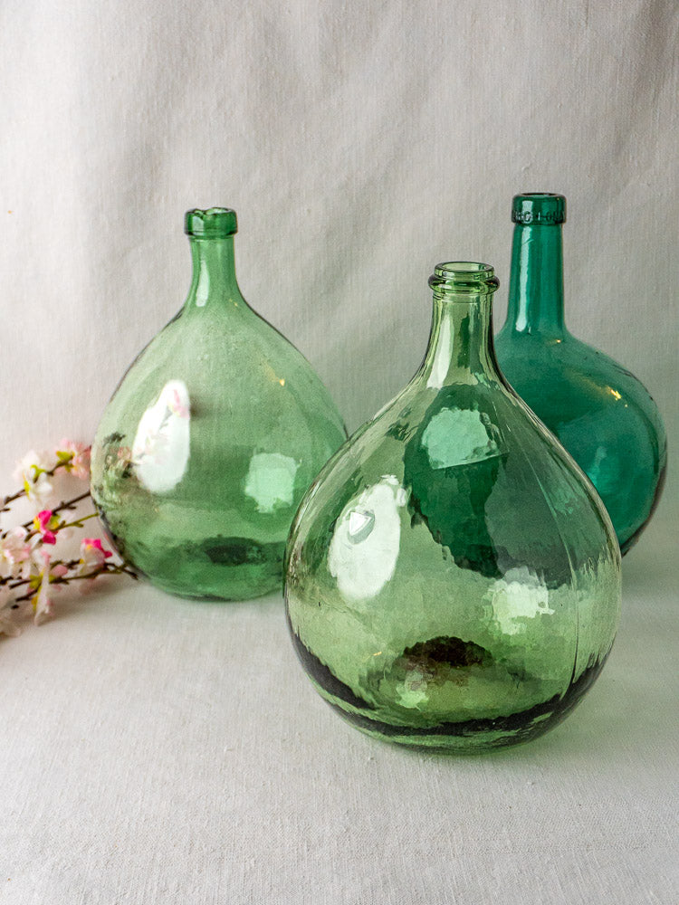 Set of three large hand-blown Spanish green glass demijohns with pink and white flowers on light fabric.