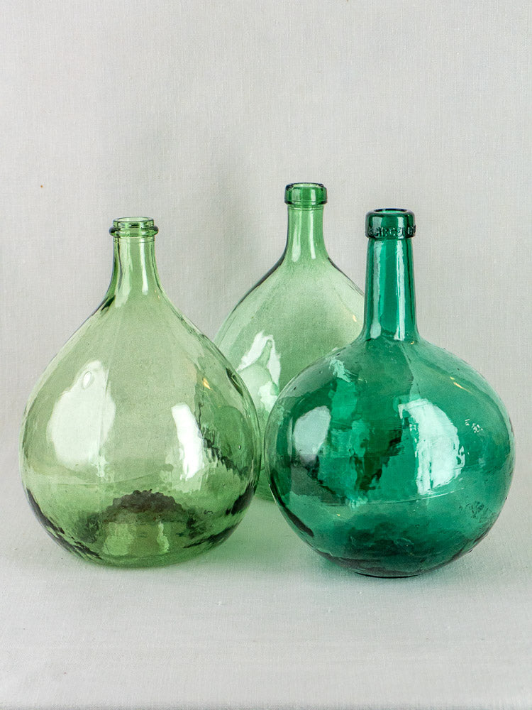 Set of three antique Spanish green glass demijohns in varying shapes on a neutral background.
